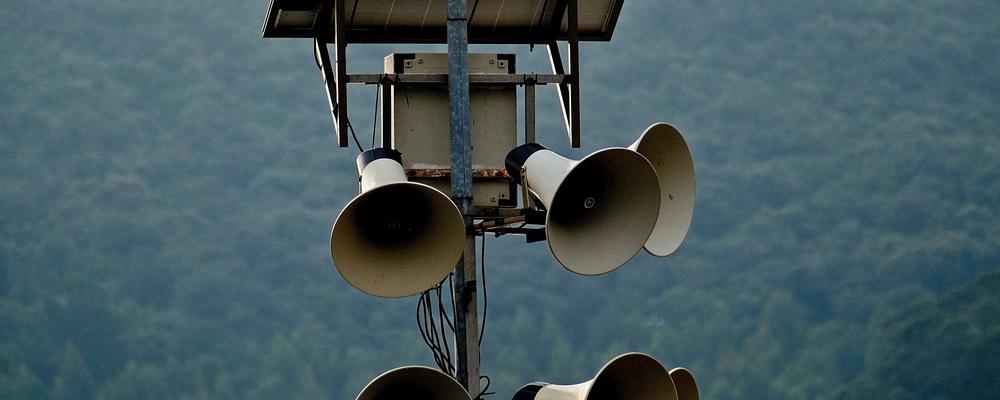 An array of loudspeakers mounted on a pole, equipped with a solar panel on top, set against a backdrop of lush green hills and a cloudy sky.