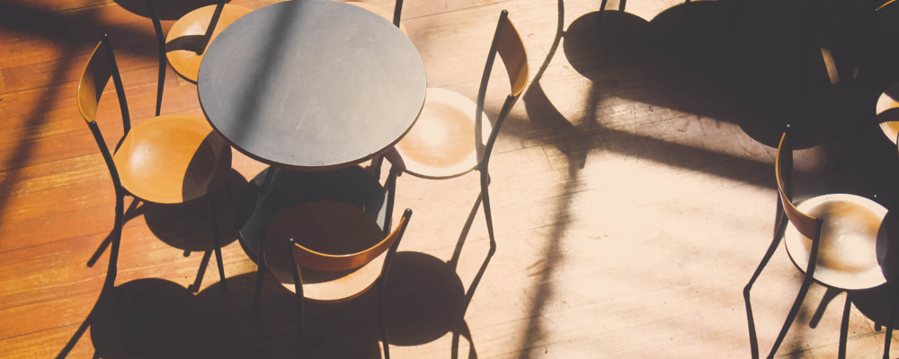 Room full of small round tables with two chairs per table. Tables casting shadows on brown wood floor.