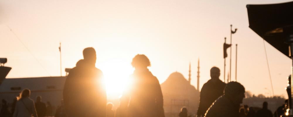 Silhouetted figures walking along a sunlit path at sunset, with a mosque dome visible in the background and a vibrant marketplace atmosphere.