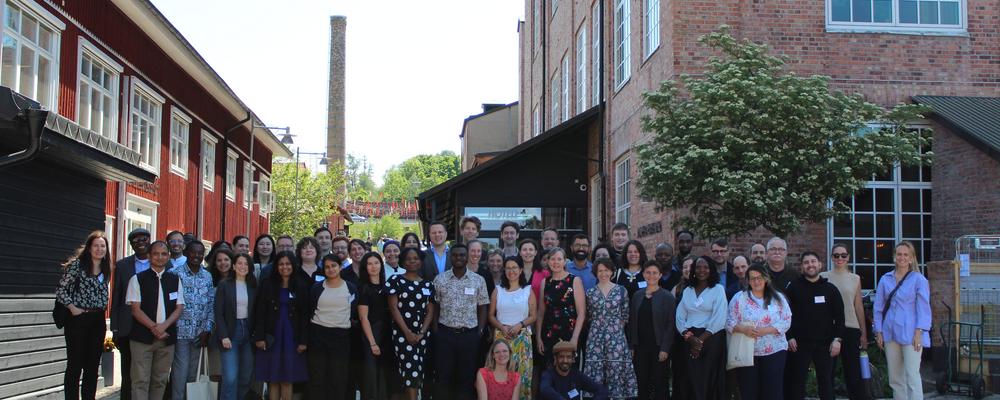 A group of 50-60 people stood between a red brick building and a red wooden building. 