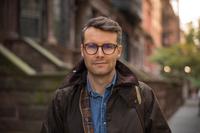 A brown-haired man in a brown jacket and blue shirt stands in a street in front of blurred red brick houses and green trees. 