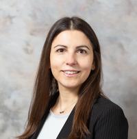 Young woman with brown hair wearing a blue blazer and white shirt in front of a grey background. 